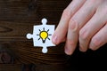 The hand holds a piece of a white puzzle with the image of a light bulb with yellow light on a wooden background. The concept of a Royalty Free Stock Photo