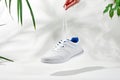 The hand holds by shoelaces white sneakers. Hand hold sporty shoes on white background with palm leaves Royalty Free Stock Photo