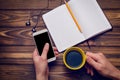 A hand holds a smartphone and a cup of coffee over a wooden table with a notebook Royalty Free Stock Photo