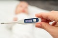 A hand holds a thermometer showing the temperature on the background of a blurred baby lying on the bed Royalty Free Stock Photo