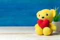 Hand make yarn red heart put on yellow teddy bear in front of white pot and green ornamental plants on wooden table and blue backg Royalty Free Stock Photo