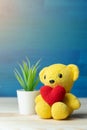 Hand make yarn red heart put on yellow teddy bear in front of white pot and green ornamental plants on wooden table and blue backg Royalty Free Stock Photo