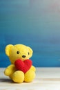 Hand make yarn red heart put on yellow teddy bear in front of white pot and green ornamental plants on wooden table and blue backg Royalty Free Stock Photo