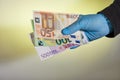 The hand of a man in a blue medical glove holds money in the form of euro bills on a yellow background. Protection against viruses