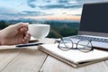 Hand man with a coffee mug Glasses on paper notebook with laptop and wood table in mountain background Royalty Free Stock Photo