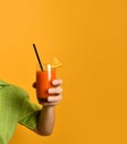 Hand of a man in green shirt holding glass of squeezed juice or cocktail with slice of lemon and tube, posing on orange background Royalty Free Stock Photo