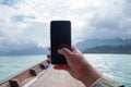 Hand of man using mobile smart phone against scenic landscape of boat view in the big river and reservoir dam with mountain and Royalty Free Stock Photo