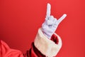 Hand of a man wearing santa claus costume and gloves over red background gesturing rock and roll symbol, showing obscene horns Royalty Free Stock Photo