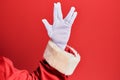 Hand of a man wearing santa claus costume and gloves over red background greeting doing vulcan salute, showing hand palm and Royalty Free Stock Photo