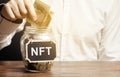 Hand puts dollar bills in glass jar with the word NFT. Non-fungible token. Digitally represented product or asset. Selling digital