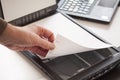 A hand puts a piece of paper on a flatbed scanner with a laptop computer in the background. Selective focus