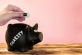 Hand putting a coin in a black piggy bank with a chalk Charity tag on a pink background, donation and charity concept Royalty Free Stock Photo