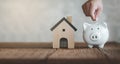 Hand putting coin on White piggy bank with wooden house on wood table and blurred copy space background. Concept for financial hom Royalty Free Stock Photo