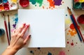 A hand rests on a blank canvas while vibrant paint spills and brushes surround a creative workspace Royalty Free Stock Photo