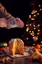 Hand Shaking Icing Sugar Over Christmas Panettone On Table Set For Meal With Tree Lights Behind Royalty Free Stock Photo