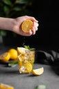 A hand squeezes juice from a lemon into a glass with lemonade with ice and mint on a dark background with citrus fruits and a Royalty Free Stock Photo