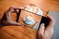 Hand taking a picture of hot latte art coffee cup with art foam on wooden table with her smart phone sitting at a coffee shop snap Royalty Free Stock Photo