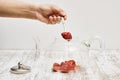 Hand taking strawberry jam with a spoon from the glass teapot on a wooden table against a white wall