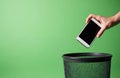 Hand throws a mobile phone into trash bin on green background. The concept of technology obsolescence. Electronic device disposal Royalty Free Stock Photo