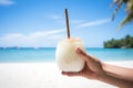 hand tipping chilled coconut, straw inserted, beach in the background