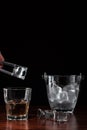 Hand with tongs holding ice-cube above a glass with whiskey, ice bucket behind the alcoholic drink on wooden table in restaurant. Royalty Free Stock Photo