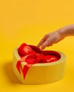 Hand tying bow on red heart-shaped box on yellow background for valentine& x27;s day