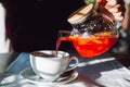 The hand of unrecognizable woman pours hot tea from glass kettle to mug sitting by table with white tablecloth. Royalty Free Stock Photo