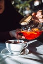 The hand of unrecognizable woman pours hot tea from glass kettle to mug sitting by table with white tablecloth. Royalty Free Stock Photo