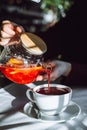 The hand of unrecognizable woman pours hot tea from glass kettle to mug sitting by table with white tablecloth. Royalty Free Stock Photo