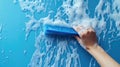 A hand using a blue squeegee to clean a soapy surface against a bright blue background Royalty Free Stock Photo