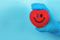 Hand wearing blue medical glove hold red smiley heart on blue background, Hands in medical gloves holding a red heart shape model