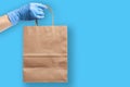Hand wearing rubber protective glove holds paper grocery bag. Grocery shopping and delivery concept during novel virus pandemic.