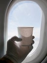 Hand in a white glove hold a white paper cup against the background of the window in the plane