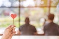 Hand of woman hold a red heart shaped candle with a blurred image of couple in the background.