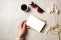Hand of woman holding blank paper card over feminine desk with hipster accessories, sunglasses, coffee cup and dry flowers. Flat Royalty Free Stock Photo