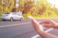 Hand of woman using smartphone and blur of her broken car parking on the road. Contacting car technician or need help concept