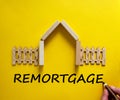 Hand writing `remortgage` on beautiful yellow background. Model of a wooden house. Male hand, wooden fence. Copy space. Business