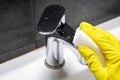 A hand in a yellow rubber glove wipes a dirty chrome metal faucet with a white melamine sponge. Cleaning in a modern bathroom