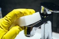 A hand in a yellow rubber glove wipes a dirty chrome metal faucet with a white melamine sponge. Cleaning in a modern bathroom