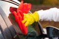 A hand in a yellow rubber glove wipes the glass of a car with a red microfiber rag from dirt on a warm autumn day. Wet cleaning. Royalty Free Stock Photo