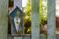 Handcrafted house shaped wooden panel with heart shape on a garden fence. Tree with bright green leaves in sunlit background.