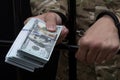 A handcuffed soldier holds out a wad of $100 paper bills from behind bars, close-up, selective focus. Concept: bribery, bail.