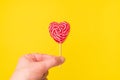 Handholds red heart-shaped lollipop candy isolated on an empty colorful yellow background. Symbol of love for Happy