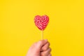 Handholds red heart-shaped lollipop candy isolated on an empty colorful yellow background. Symbol of love for Happy Women`s,