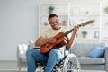 Handicapped black guy in wheelchair playing guitar and smiling at living room. Stay home activities Royalty Free Stock Photo