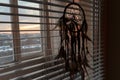 Handmade dreamcatcher hanging by the window with blinds in sunset twilight. Black silhouette of traditional magic amulet for dream