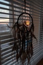 Handmade dreamcatcher hanging by the window with blinds in sunset twilight. Black silhouette of traditional magic amulet for dream