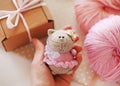 Handmade knitted toy. Knitted cat in a pink dress and bow in female hand on a background of gift box. Birthday gift idea