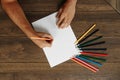 Hands of the artist close-up, holds a pencil over a blank sheet of paper near scattered colored pencils on a brown wooden table. Royalty Free Stock Photo
