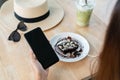 Hands of Asian woman using smartphone while eating brownie cake with iced Matcha latte, beach hat and sunglasses on wooden table